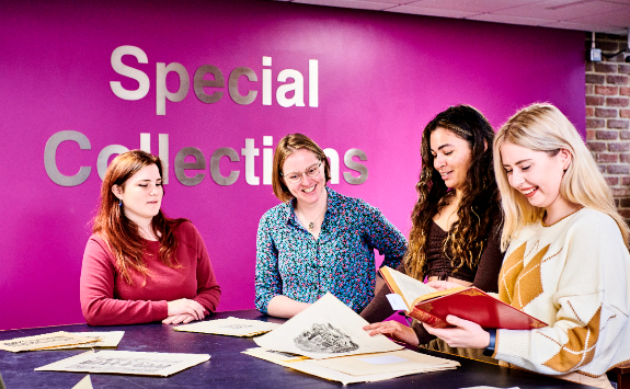 A group of 3 students and a teacher in the middle, stood around a desk looking and smiling at Special Collections paper material, whilst stood in front of a purple wall with 'Special Collections' lettering